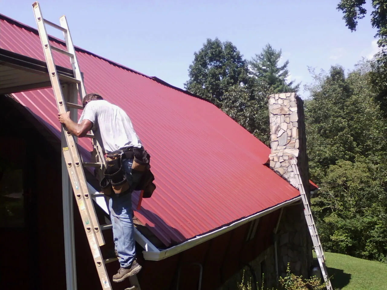 Roofer on a ladder beside a red metal roof and stone chimney — Soffit & Fascia Repair by Roofing Brothers in Goldsboro
